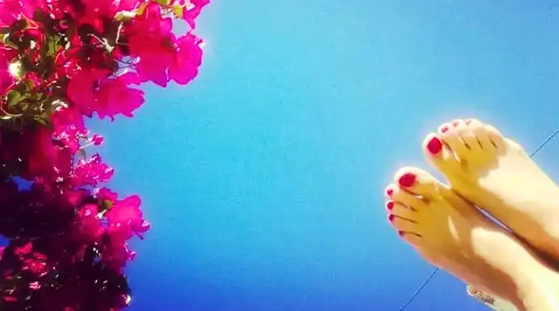 Pies con uñas pintadas de rosado, y flores rosadas, con cielo azul de fondo.